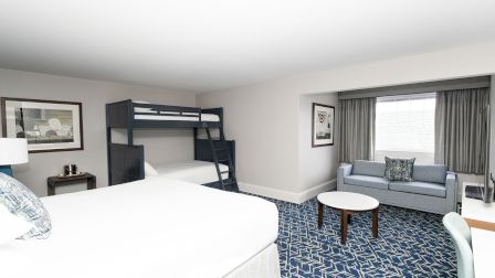 A bright hotel room with a queen bed, bunk bed, blue patterned carpet, a small sofa, coffee table, and a window with gray curtains, ending with period.