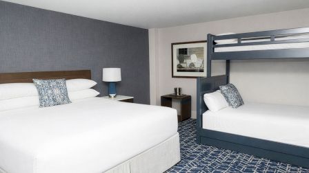 A hotel room with a king bed and a bunk bed setup, blue and white decor, nightstand, lamp, framed art, and patterned carpet.