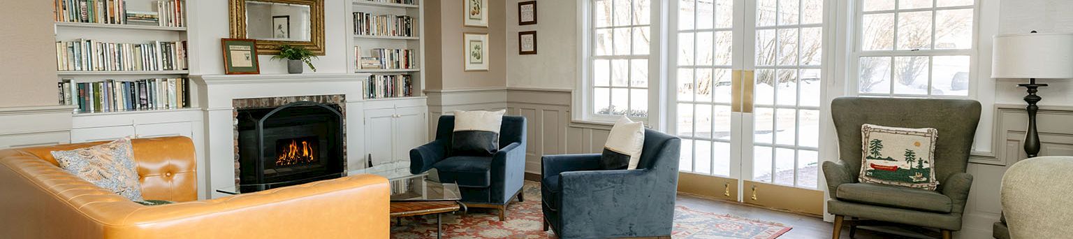 A spacious living room with a fireplace, built-in bookshelves, several chairs and a sofa, large windows, and a patterned area rug.