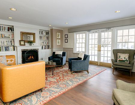 A spacious living room with a fireplace, built-in bookshelves, several chairs and a sofa, large windows, and a patterned area rug.