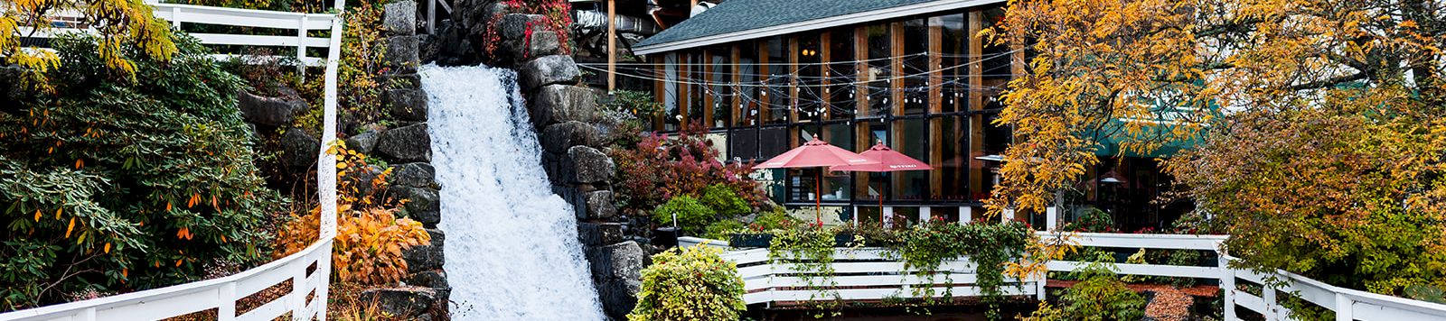 A scenic riverside resort with a cascading waterfall, white buildings, wooden railings, autumn foliage, and a patio with red umbrella.