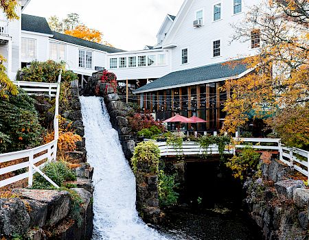 A scenic riverside resort with a cascading waterfall, white buildings, wooden railings, autumn foliage, and a patio with red umbrella.
