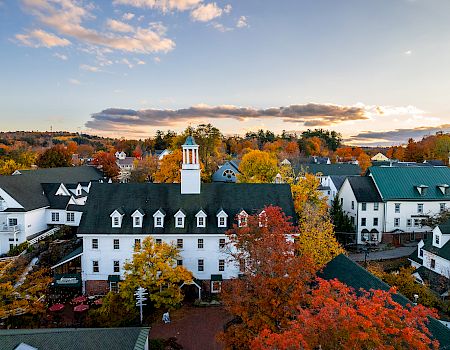 An aerial view of a small town with white buildings, autumn trees in orange and red, and a central clock tower.