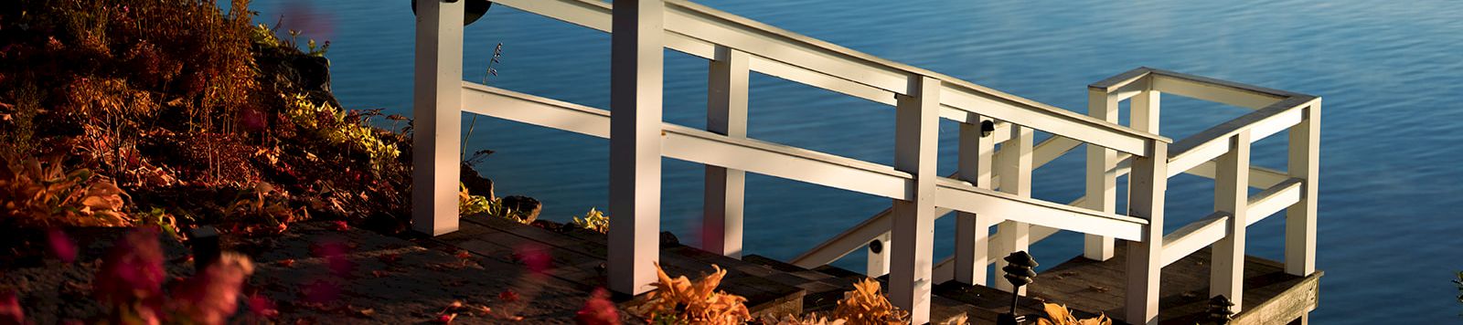 A tranquil lakeside scene with a wooden viewing platform extending over calm blue waters, mist rising from the surface, and autumn foliage framing the shore.