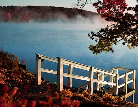 A tranquil lakeside scene with a wooden viewing platform extending over calm blue waters, mist rising from the surface, and autumn foliage framing the shore.