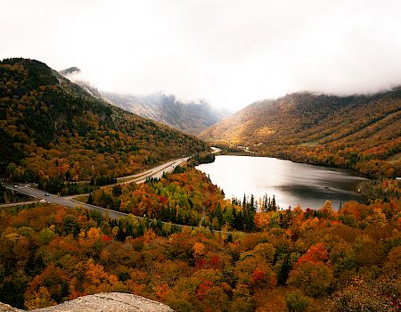 A winding mountain road curves through a colorful autumn valley with a tranquil lake, surrounded by trees in orange, red, and gold, under cloudy skies.