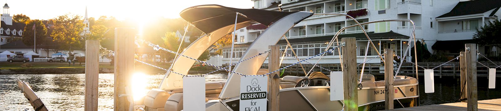 A sunny marina at dawn with a wooden dock, a white boat, and a large white multi-story hotel or resort in the background.