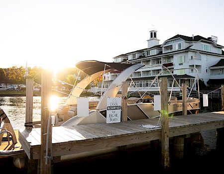 A sunny marina at dawn with a wooden dock, a white boat, and a large white multi-story hotel or resort in the background.