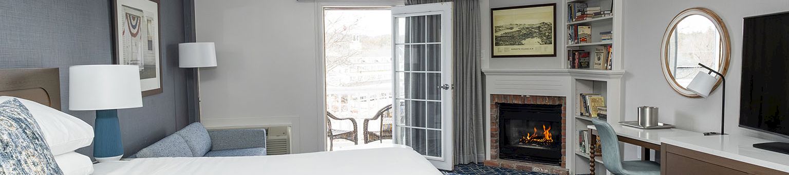 A modern hotel room with a large bed, blue accents, a fireplace, desk, balcony doors, and a flat-screen TV, all in a clean, airy layout.