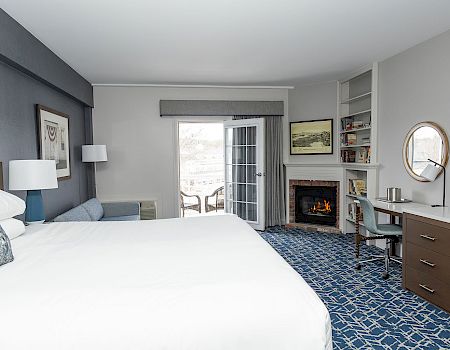 A modern hotel room with a large bed, blue accents, a fireplace, desk, balcony doors, and a flat-screen TV, all in a clean, airy layout.