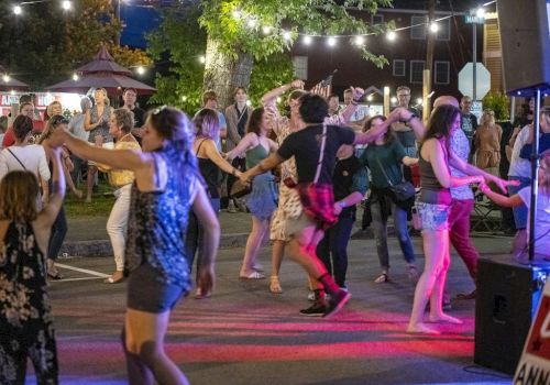 A lively outdoor dance party with people twirling, swapping partners, and enjoying string lights under a night market vibe.