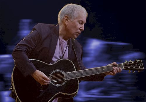 A man playing a black acoustic guitar on stage, blurred blue lights in the background.