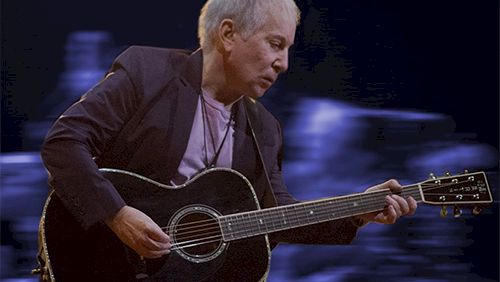 A man playing a black acoustic guitar on stage, blurred blue lights in the background.