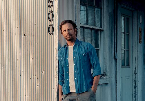 A casually dressed man leans against a weathered storefront with peeling blue doors, wearing a blue shirt over a white tee and gray jeans.