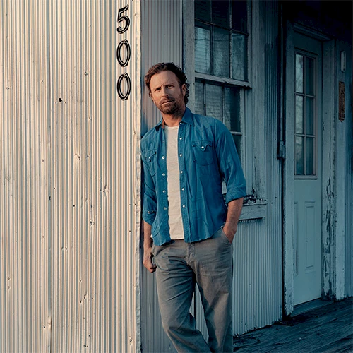 A casually dressed man leans against a weathered storefront with peeling blue doors, wearing a blue shirt over a white tee and gray jeans.