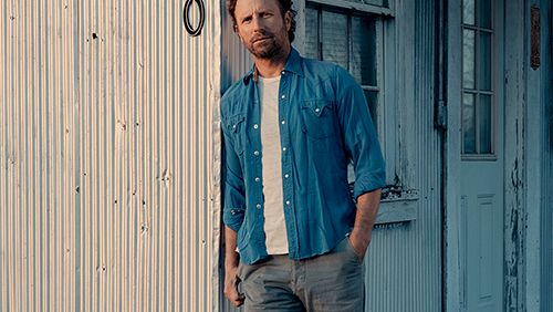 A casually dressed man leans against a weathered storefront with peeling blue doors, wearing a blue shirt over a white tee and gray jeans.