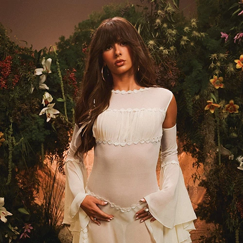 A woman in a sheer white dress with puffed sleeves stands in a garden setting with flowers and greenery around her. Ending sentence with a period.