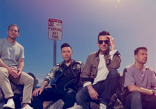 Four men pose on rocks by a &ldquo;No Parking Any Time&rdquo; sign, casual 90s vibe, sunny sky, relaxed group photo.