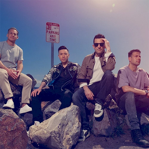 Four men pose on rocks by a &ldquo;No Parking Any Time&rdquo; sign, casual 90s vibe, sunny sky, relaxed group photo.