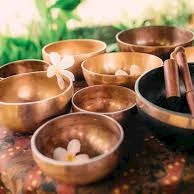 A set of copper singing bowls on a patterned cloth, with a few wooden mallets and delicate flowers, creating a calming altar-like scene.