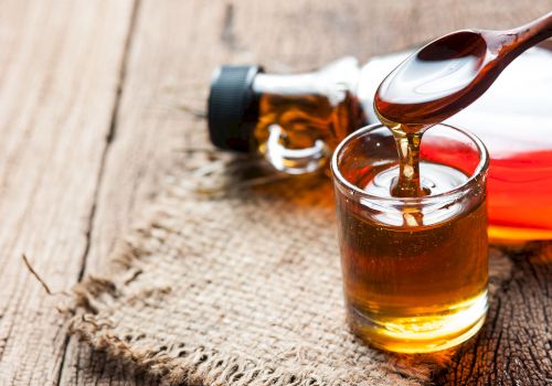 A small glass of maple syrup with a wooden spoon; bottle and wood surface in the background.