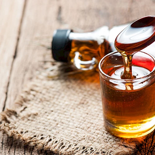 A small glass of maple syrup with a wooden spoon; bottle and wood surface in the background.