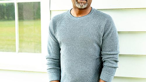 A man stands outside in a light gray sweater and jeans, hands in pockets, beside a cream-colored house with a window.