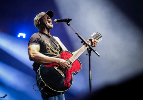 A male musician wearing a cap, playing a red acoustic-electric guitar and singing into a microphone on stage with blue spotlight and smoky backdrop.