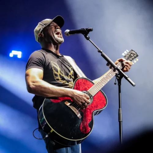 A male musician wearing a cap, playing a red acoustic-electric guitar and singing into a microphone on stage with blue spotlight and smoky backdrop.