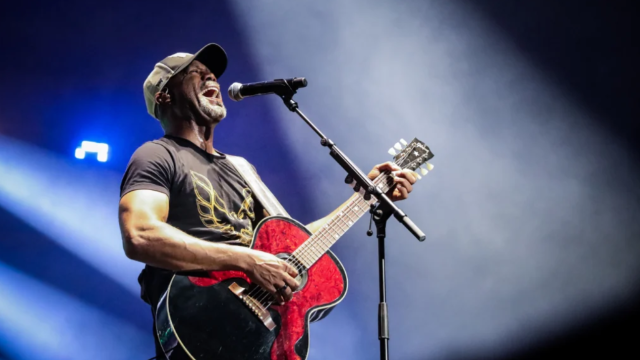 A male musician wearing a cap, playing a red acoustic-electric guitar and singing into a microphone on stage with blue spotlight and smoky backdrop.