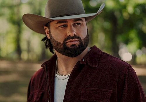 A man with a beard wearing a wide-brimmed hat and burgundy shirt stands outdoors in a leafy park.