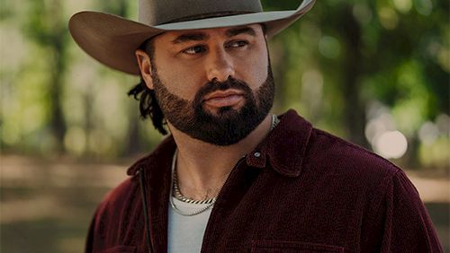 A man with a beard wearing a wide-brimmed hat and burgundy shirt stands outdoors in a leafy park.
