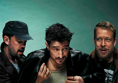 Three men pose together, one in a green shirt center, glasses off, black jackets, teal backdrop.