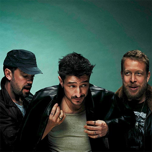 Three men pose together, one in a green shirt center, glasses off, black jackets, teal backdrop.