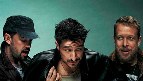 Three men pose together, one in a green shirt center, glasses off, black jackets, teal backdrop.