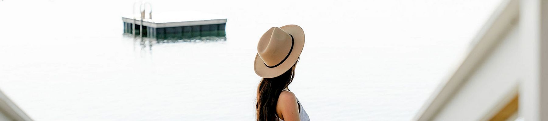 A woman in a hat stands on a white pier railing overlooking water, wearing a white top and denim shorts.
