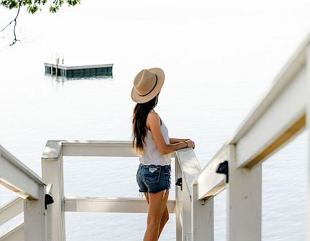 A woman in a hat stands on a white pier railing overlooking water, wearing a white top and denim shorts.