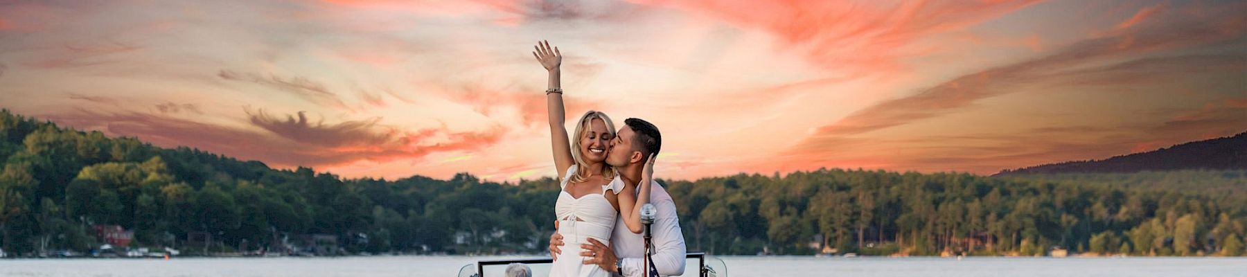 Two people in stylish outfits cheer on a wooden boat named "Mill Falls" on a calm lake at sunset, with US flag waving.