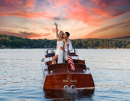 Two people in stylish outfits cheer on a wooden boat named "Mill Falls" on a calm lake at sunset, with US flag waving.