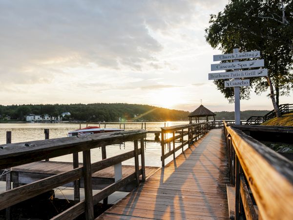 A wooden dock extends toward the sunset over calm water, with signposts and trees on the right and a sheltered gazebo at the end.