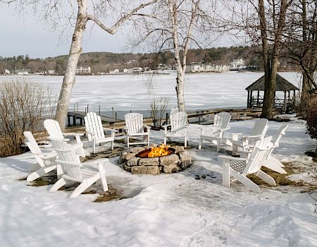 A snowy lakeside scene with white Adirondack chairs arranged in a circle around a fire pit, bare trees, and a cozy winter landscape.