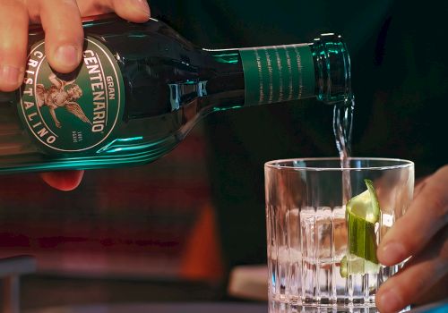 Pouring green bottled beverage into a glass with lime wedge, hands visible; close-up bar scene, condensation on bottle, dark background ending this sentence.