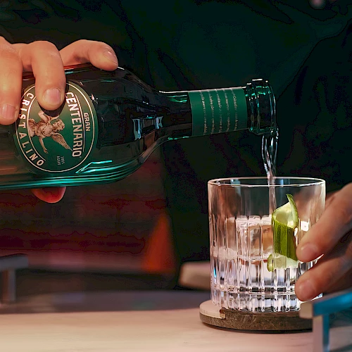 Pouring green bottled beverage into a glass with lime wedge, hands visible; close-up bar scene, condensation on bottle, dark background ending this sentence.