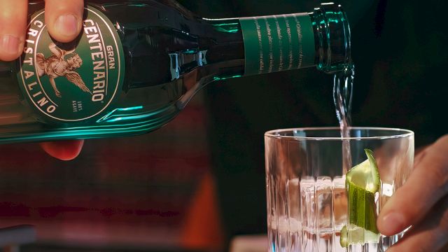 Pouring green bottled beverage into a glass with lime wedge, hands visible; close-up bar scene, condensation on bottle, dark background ending this sentence.