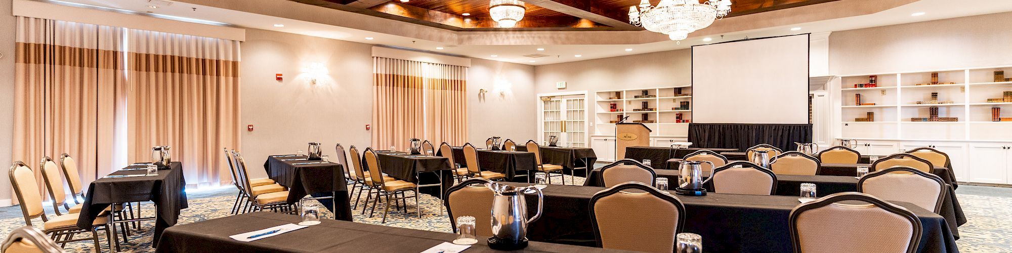 This image shows a conference room setup with tables and chairs, chandeliers, and a projector screen, creating an elegant and professional atmosphere.