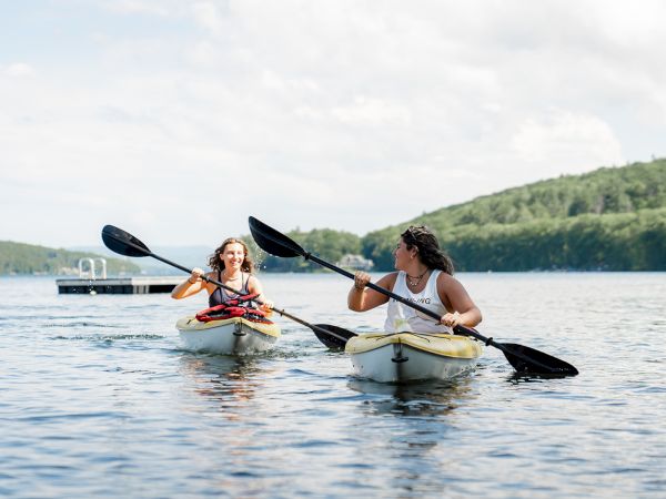 Two people kayaking on a calm lake, paddling side by side with green hills in the background.