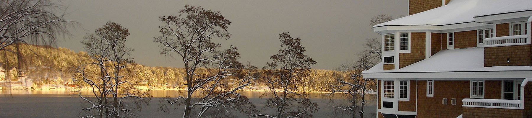 Snowy landscape with a lake, leafless trees, and a brick building with a white roof and a clock tower in the distance, calm winter scene.