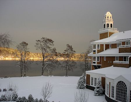 Snowy landscape with a lake, leafless trees, and a brick building with a white roof and a clock tower in the distance, calm winter scene.