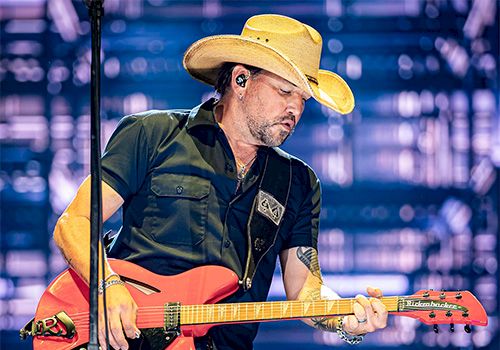 A man wearing a cowboy hat plays a red electric guitar on stage, performing with intensity under bright backstage lights.