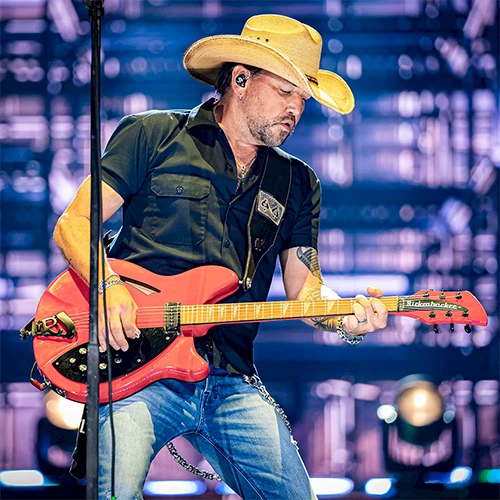 A man wearing a cowboy hat plays a red electric guitar on stage, performing with intensity under bright backstage lights.